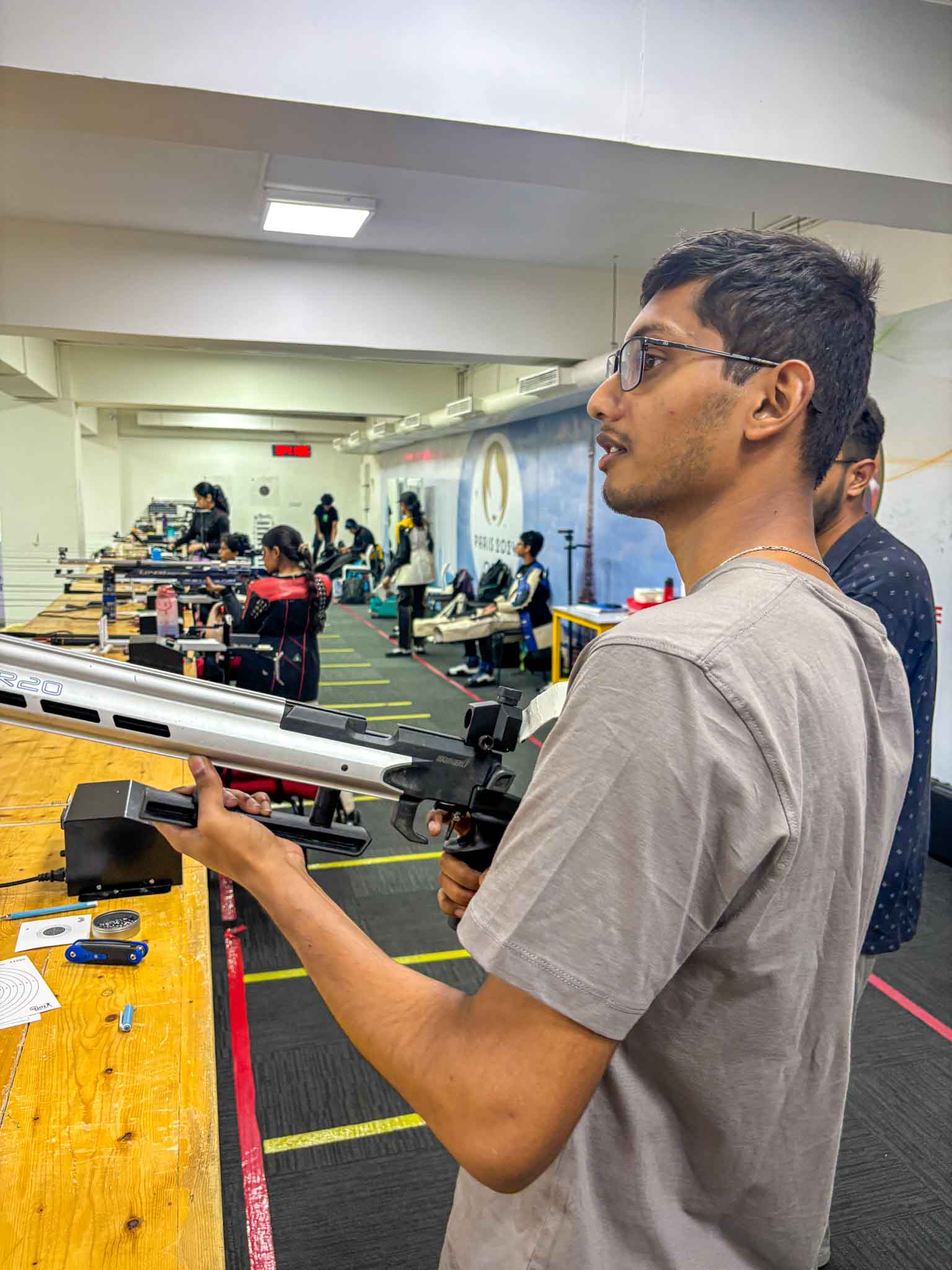 A shooting instructor adjusting a male participant's rifle sight at an indoor shooting gallery.